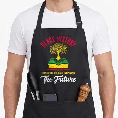 Honoring african apron - Yellow tree growing from open book with red BLACK HISTORY text and white script — shown on a black cotton canvas apron against a white background