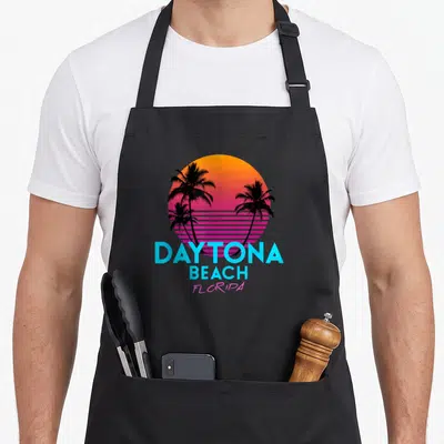 Daytona 80s apron - Retro gradient sun with black palm trees and cyan DAYTONA BEACH FLORIDA text — shown on a black cotton canvas apron against a white background