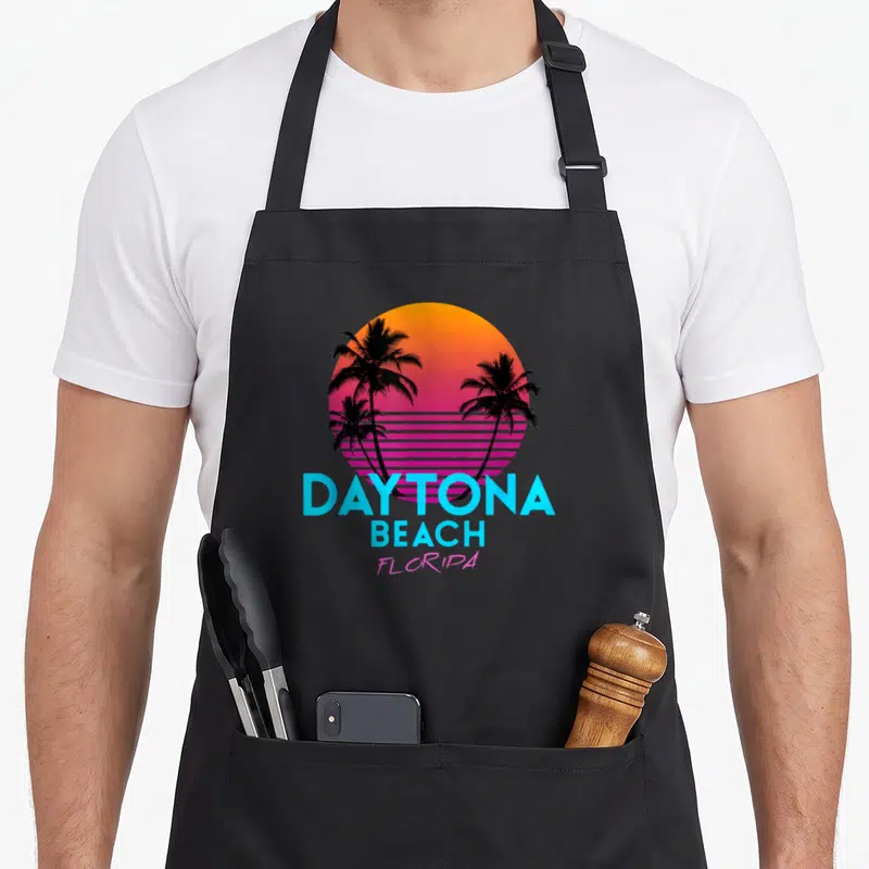 Daytona 80s apron - Retro gradient sun with black palm trees and cyan DAYTONA BEACH FLORIDA text — shown on a black cotton canvas apron against a white background