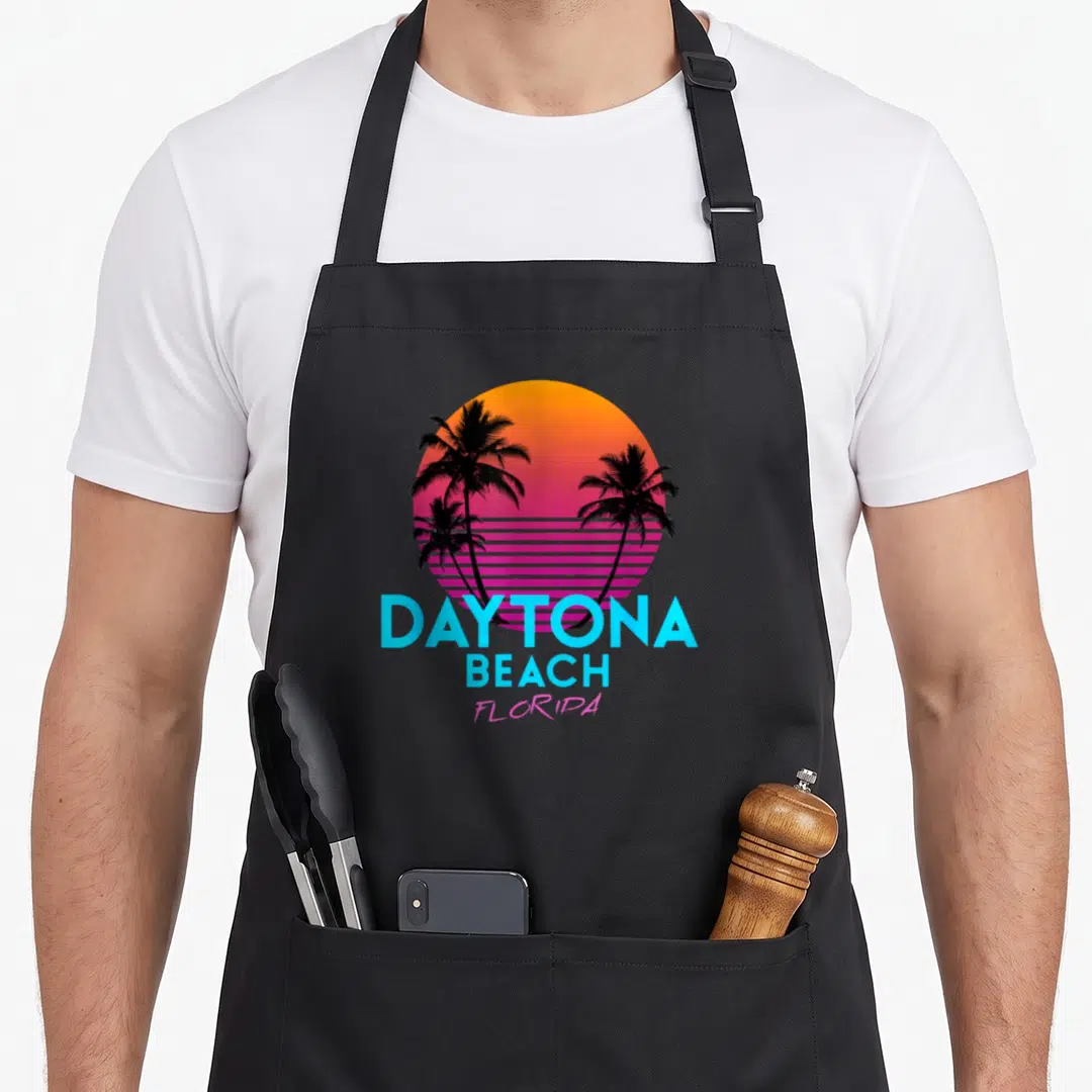Daytona 80s apron - Retro gradient sun with black palm trees and cyan DAYTONA BEACH FLORIDA text — shown on a black cotton canvas apron against a white background