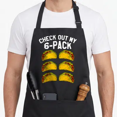 Fitness taco apron - CHECK OUT MY 6-PACK text above six cartoon tacos with yellow shells and red filling — shown on a black cotton canvas apron against a white background