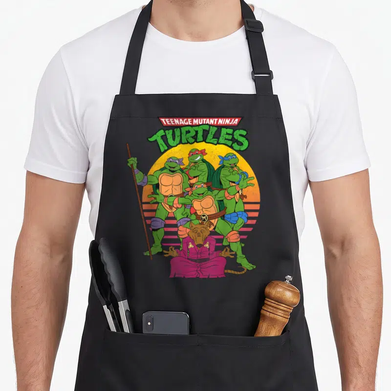 Sun teenage cooking aprons - Four turtles and a rat sitting cross-legged in front of a yellow striped circle — shown on a black cotton canvas apron against a white background