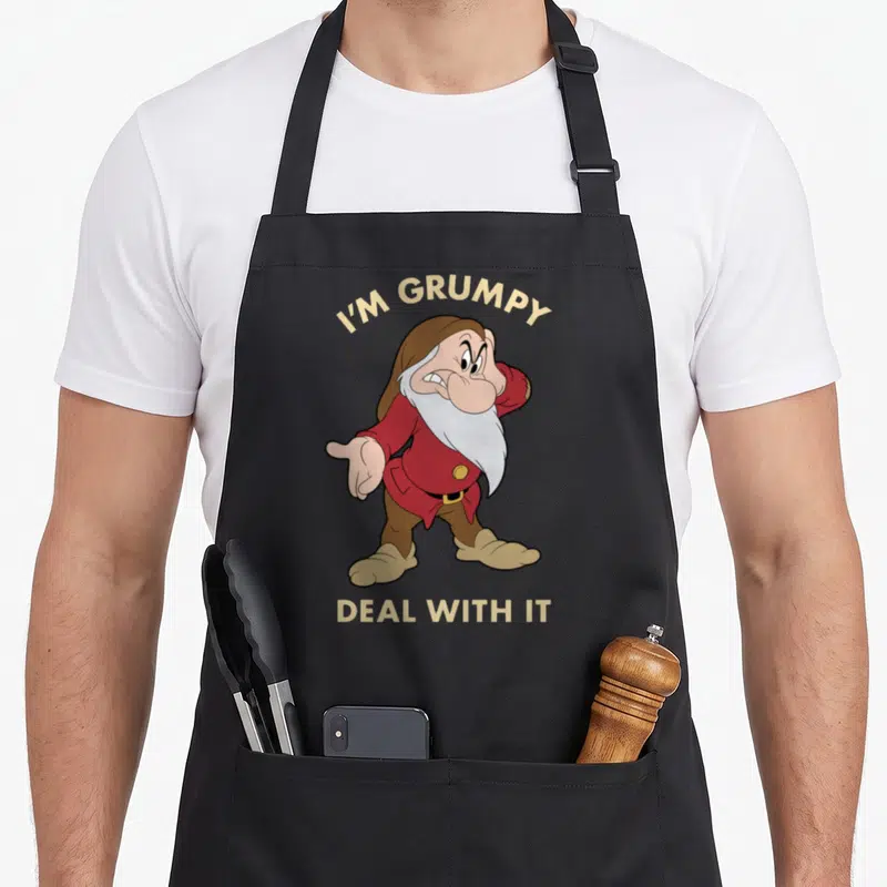 Deal snow white apron - Cartoon dwarf in red coat with text I'M GRUMPY DEAL WITH IT arched above and straight below — shown on a black cotton canvas apron against a white background