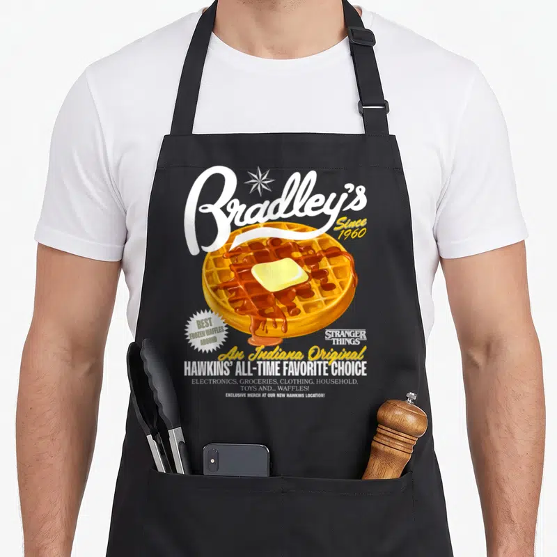 Season frozen apron - Retro waffle with butter and syrup under white Bradley's script and yellow Indiana Original text — shown on a black cotton canvas apron against a white background