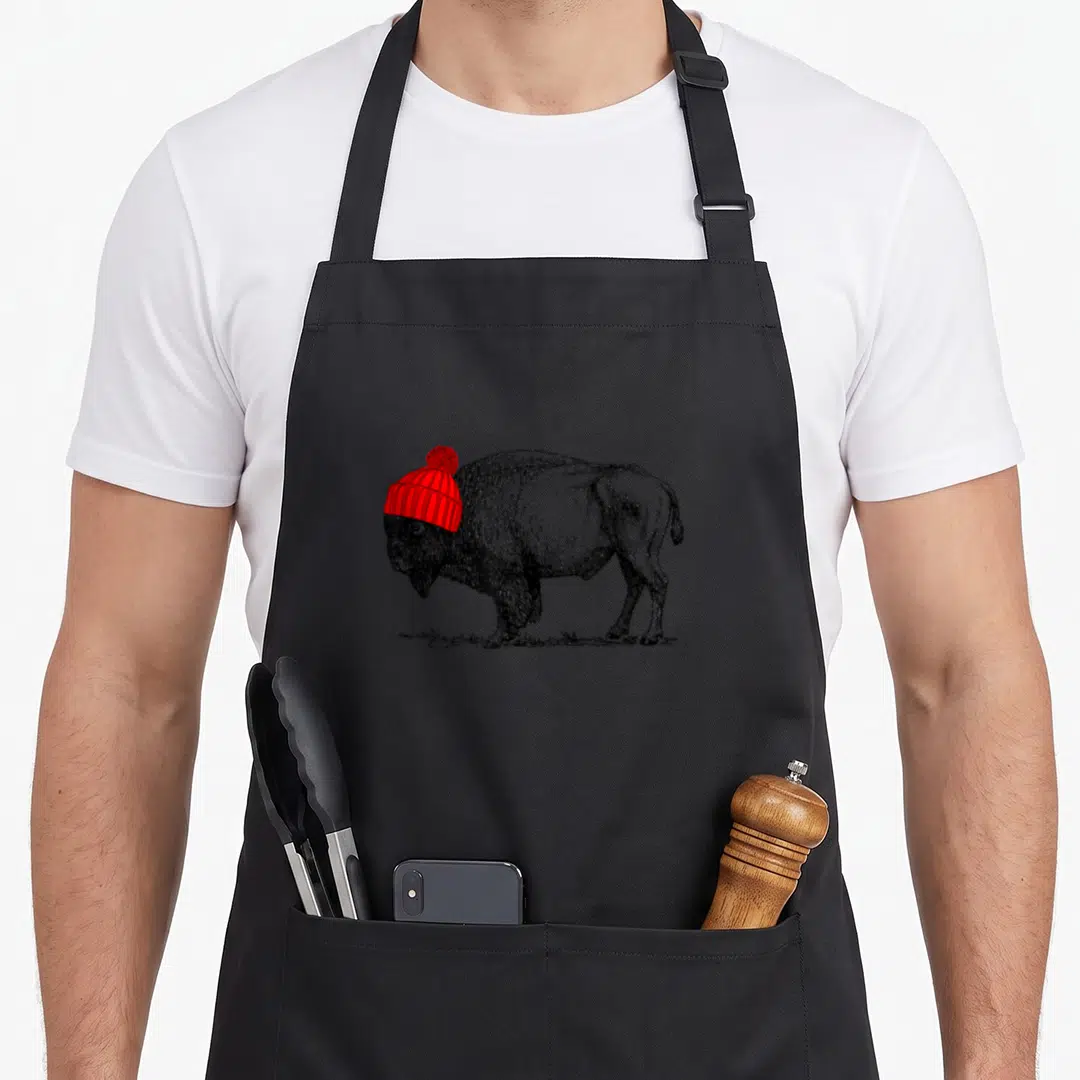 Beanie buffalo apron - Black and white bison sketch wearing a red ribbed beanie with a pom-pom on top — shown on a black cotton canvas apron against a white background