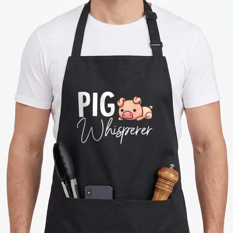 Whisperer pig lovers apron - Cartoon pink pig lying down next to white PIG Whisperer text — shown on a black cotton canvas apron against a white background
