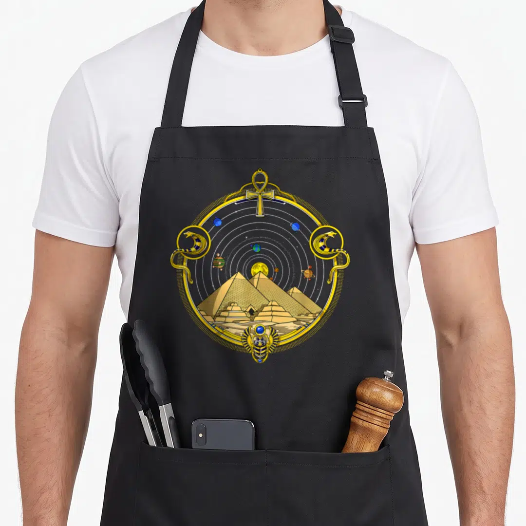 Pyramid egyptian apron - Circular gold frame with pyramids, an ankh, two cobras, and a winged scarab in cartoon style — shown on a black cotton canvas apron against a white background
