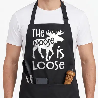 Loose elk apron - White moose silhouette with integrated text reading THE MOOSE is LOOSE in distressed font — shown on a black cotton canvas apron against a white background