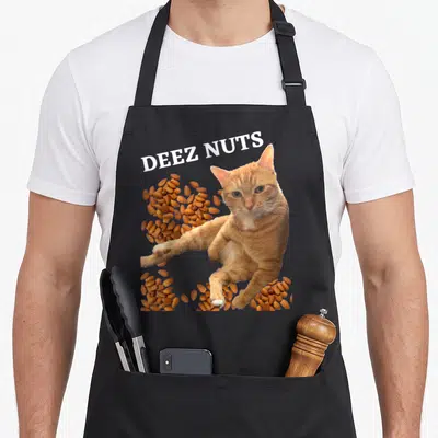 Cat joke apron - Deez Nuts text over orange cat surrounded by brown almond clusters — shown on a black cotton canvas apron against a white background