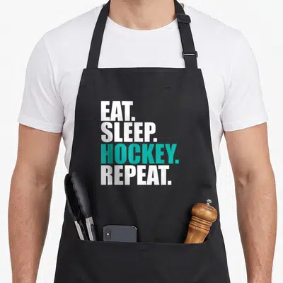 Eat hockey dad apron - Stacked white and teal text reading EAT SLEEP HOCKEY REPEAT on black fabric — shown on a black cotton canvas apron against a white background