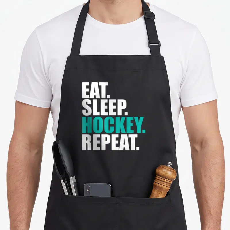 Eat hockey dad apron - Stacked white and teal text reading EAT SLEEP HOCKEY REPEAT on black fabric — shown on a black cotton canvas apron against a white background