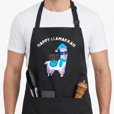Cklich hanukkah apron - Cartoon llama with sunglasses and menorah under Happy Llamakkah text — shown on a black cotton canvas apron against a white background