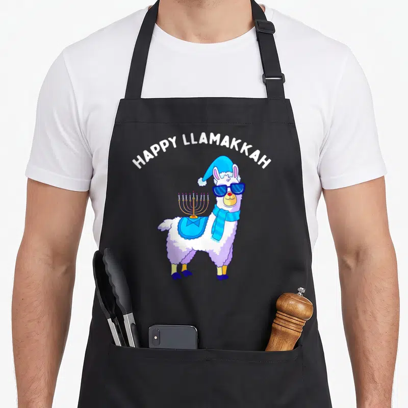 Cklich hanukkah apron - Cartoon llama with sunglasses and menorah under Happy Llamakkah text — shown on a black cotton canvas apron against a white background