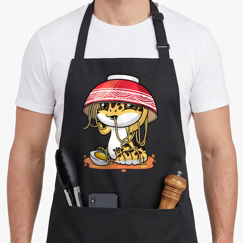 Kawaii leopard apron - Cartoon leopard in red bowl hat with yellow noodles and halved egg in orange soup — shown on a black cotton canvas apron against a white background