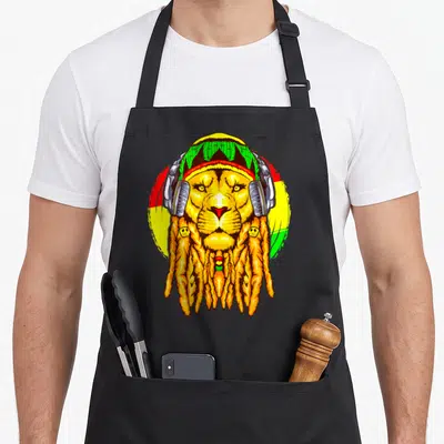 Horoscope lion apron - Cartoon lion with Rasta hat, headphones, and orange dreadlocks against red yellow green circle — shown on a black cotton canvas apron against a white background