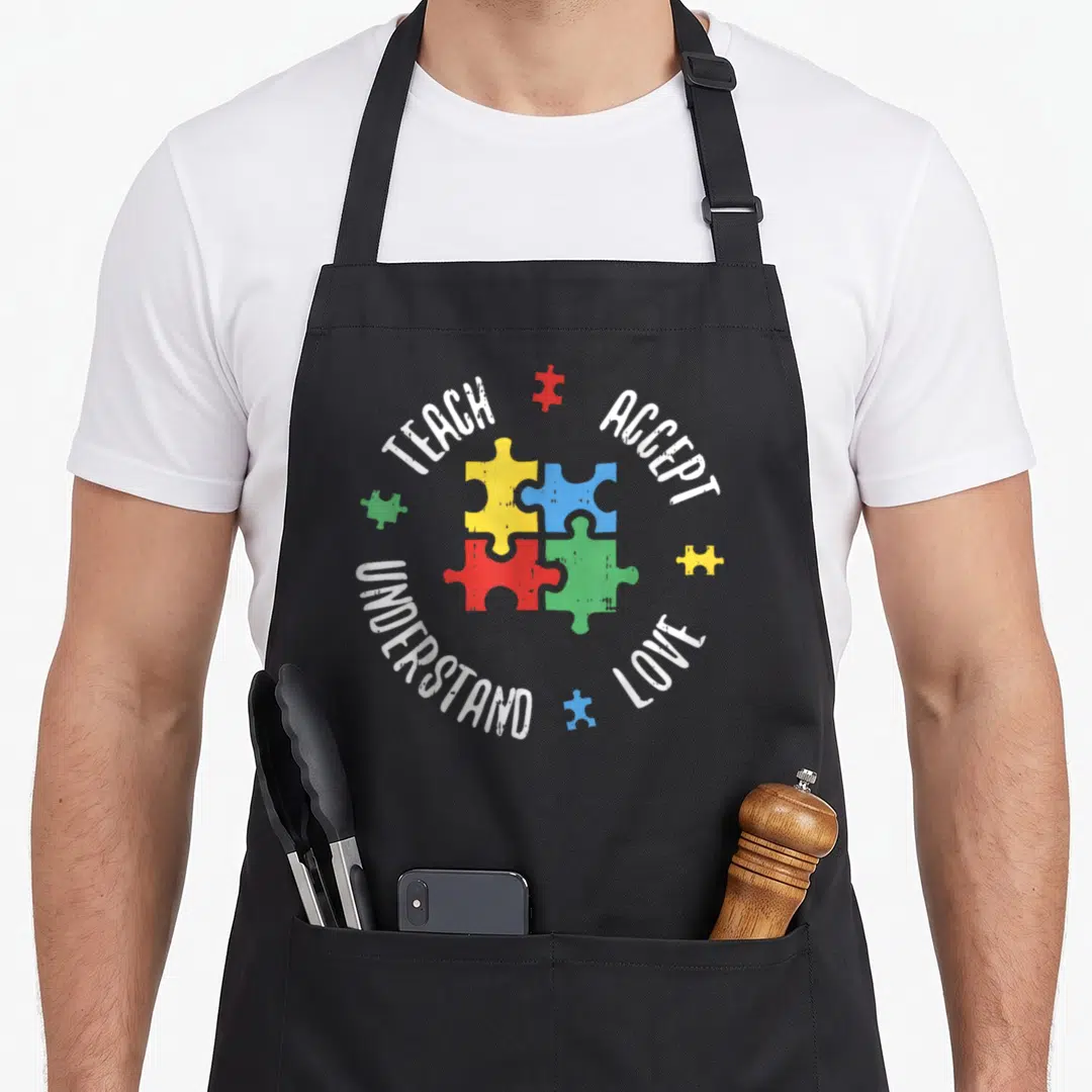 Autism teacher art apron - Teach Accept Love Understand text around yellow blue red green puzzle pieces distressed print — shown on a black cotton canvas apron against a white background