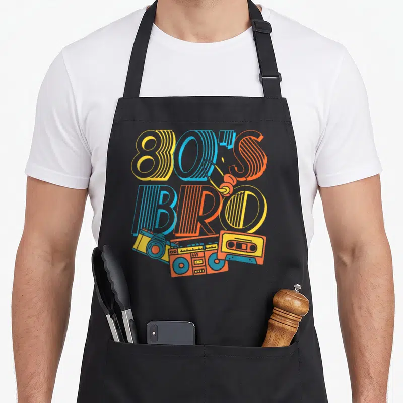 Bro cassette 80s apron - Yellow blue orange 80S BRO text with headphones camera boombox and cassette tape — shown on a black cotton canvas apron against a white background