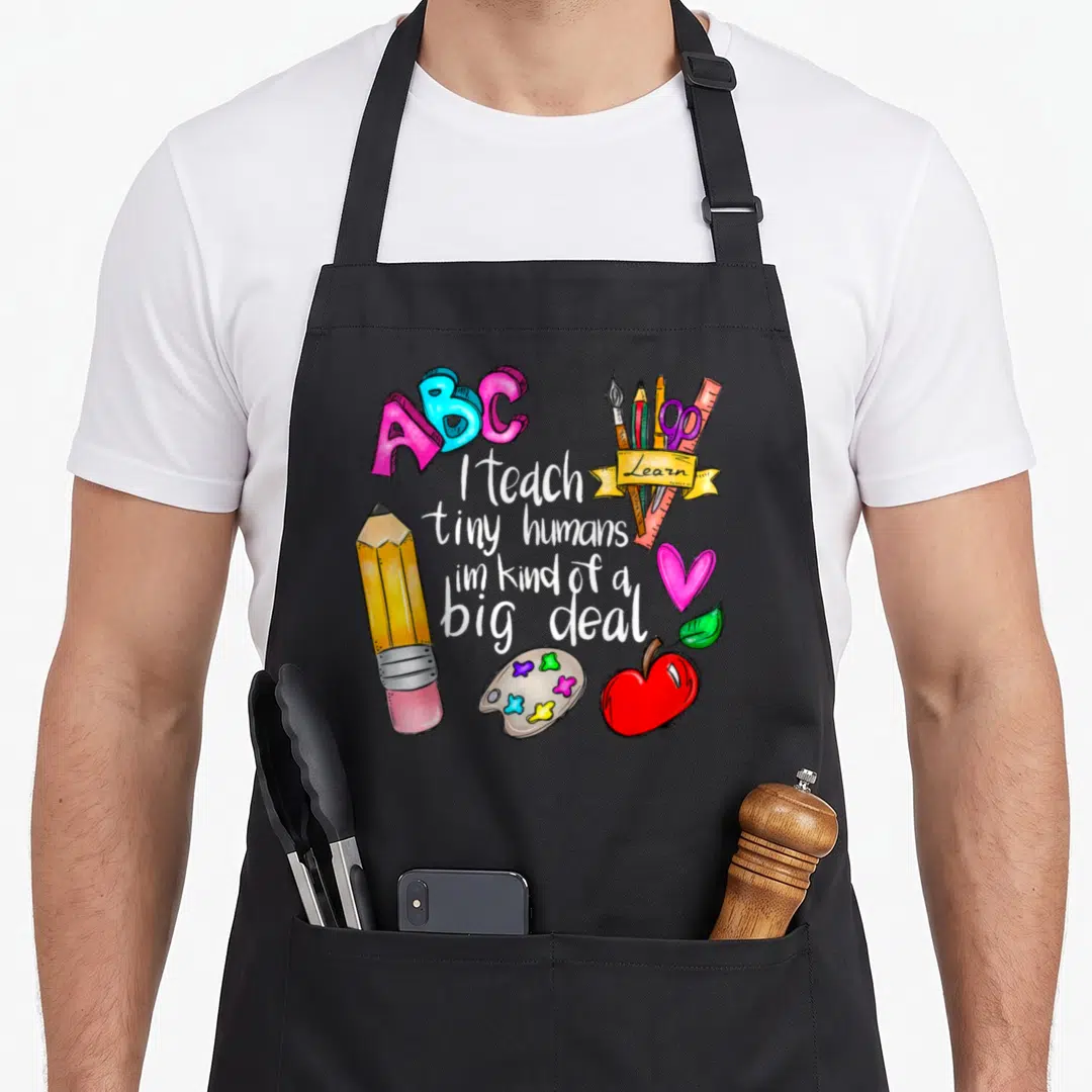 Tiny teacher apron - I teach tiny humans text surrounded by ABCs, pencil, apple, palette, and tools in pink blue yellow — shown on a black cotton canvas apron against a white background