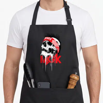 Skull machine apron - Dripping cartoon skull with red bandana reading ST 19XX and red MGK text behind it — shown on a black cotton canvas apron against a white background