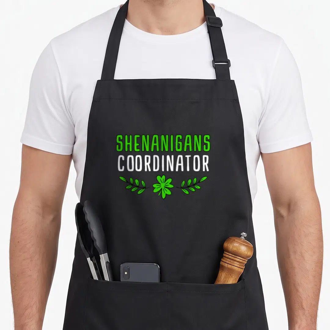 Coordinator st patrick day aprons - Green and white Shenanigans Coordinator text above a shamrock and two leafy stems — shown on a black cotton canvas apron against a white background