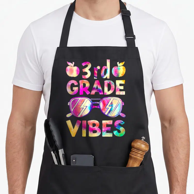 3rd tie-dye apron - Tie-dye 3rd GRADE VIBES text with sunglasses and two apples in pink blue yellow purple — shown on a black cotton canvas apron against a white background