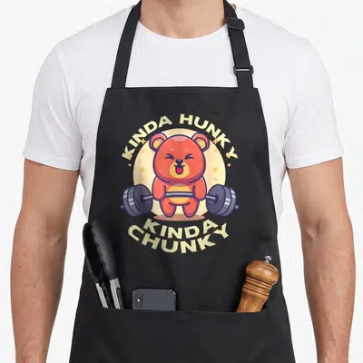Kinda loose funny apron - Red cartoon bear holding barbell with arched yellow text KINDA HUNKY KINDA CHUNKY — shown on a black cotton canvas apron against a white background