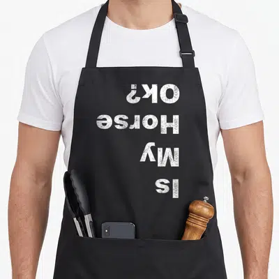 Vintage riding horse apron - White text reading Is My Horse OK printed upside down on black fabric — shown on a black cotton canvas apron against a white background