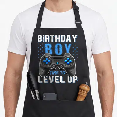 Level gamer birthday apron - Black game controller with blue buttons surrounded by blue plus signs and white and blue — shown on a black cotton canvas apron against a white background