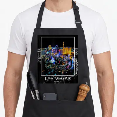 Las lv apron - Framed night photo of Las Vegas skyline with white border and location text below — shown on a black cotton canvas apron against a white background