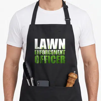 Lawn grass cutting apron - LAWN ENFORCEMENT OFFICER text filled with green grass photo and white top line — shown on a black cotton canvas apron against a white background