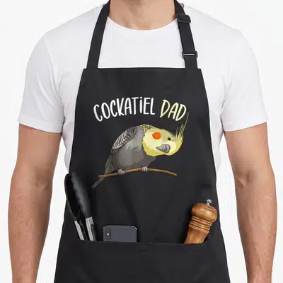Vintage cockatiel bird apron - Cartoon cockatiel perched on brown branch under arched COCKATIEL DAD text in white — shown on a black cotton canvas apron against a white background