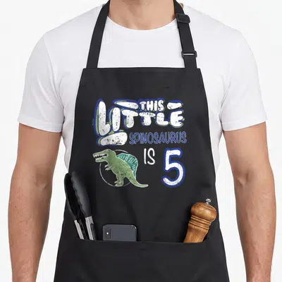 Spinosaurus 5th dinosaur apron - Green spinosaurus in white circle with blue and white text reading THIS LITTLE — shown on a black cotton canvas apron against a white background