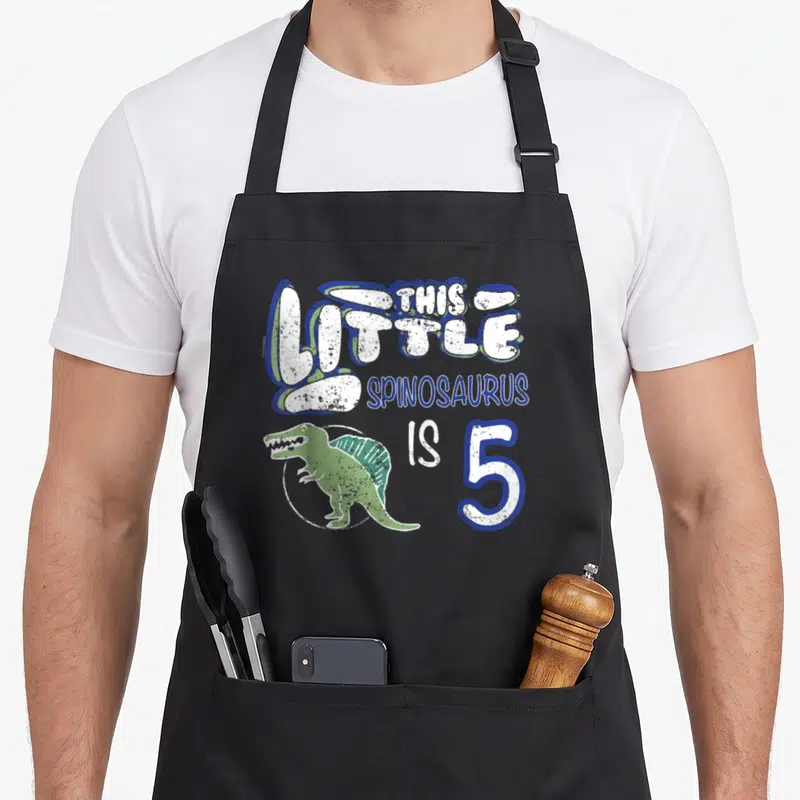 Spinosaurus 5th dinosaur apron - Green spinosaurus in white circle with blue and white text reading THIS LITTLE — shown on a black cotton canvas apron against a white background