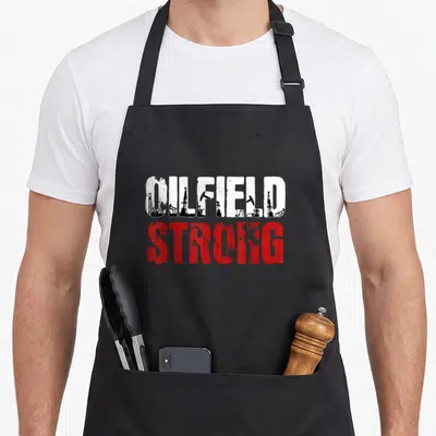 Oilfield oil apron - OILFIELD STRONG text with oil rig silhouettes inside white letters and red stacked text — shown on a black cotton canvas apron against a white background