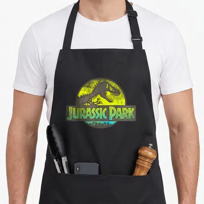 Safari novelty apron - T-Rex skeleton in yellow-green circle with JURASSIC PARK banner and palm trees — shown on a black cotton canvas apron against a white background