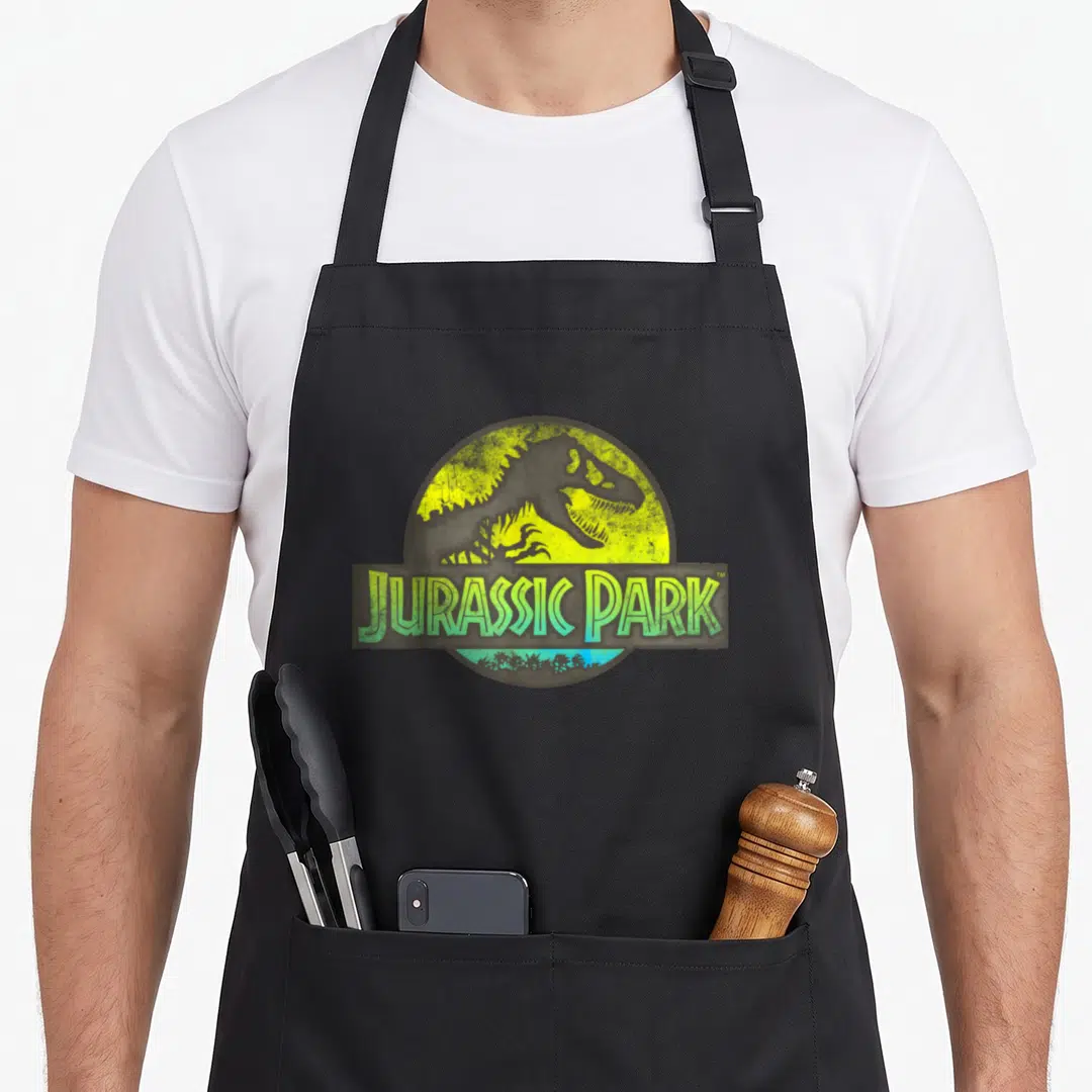 Safari novelty apron - T-Rex skeleton in yellow-green circle with JURASSIC PARK banner and palm trees — shown on a black cotton canvas apron against a white background