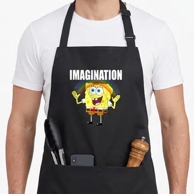 Imagination rainbow apron - SpongeBob SquarePants standing under a rainbow arc with white IMAGINATION text above — shown on a black cotton canvas apron against a white background