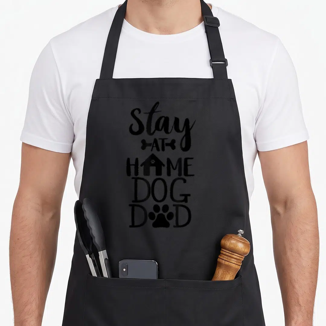 Cute dog stay at home dad apron - Stacked black text Stay AT HOME DOG DAD with bone house and paw print icons — shown on a black cotton canvas apron against a white background