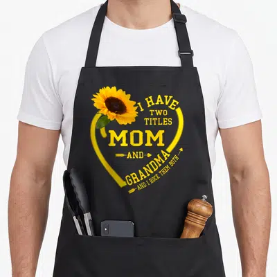 Title grandma mother's day aprons - Sunflower inside yellow heart with text I HAVE TWO TITLES MOM AND GRANDMA AND I — shown on a black cotton canvas apron against a white background