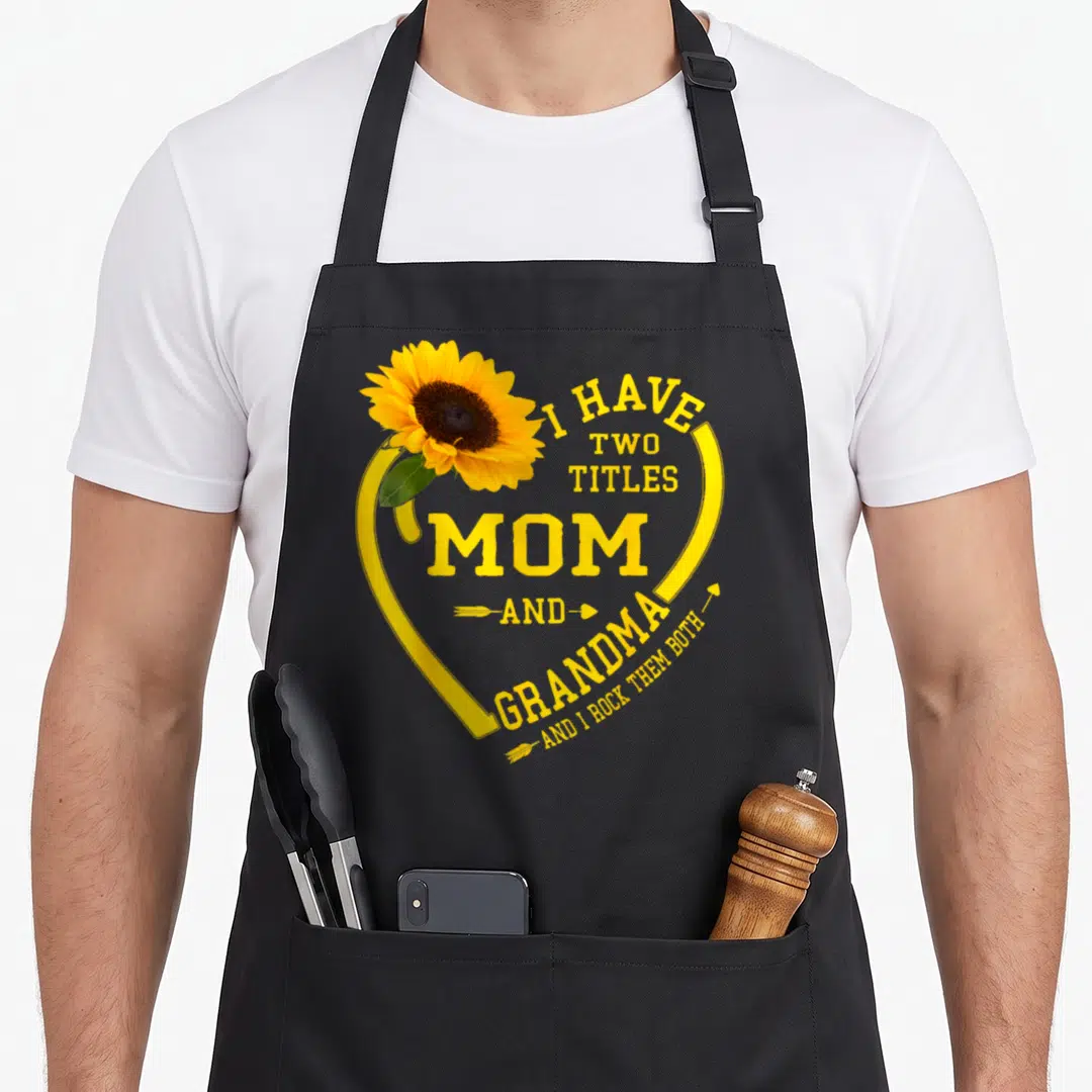 Title grandma mother's day aprons - Sunflower inside yellow heart with text I HAVE TWO TITLES MOM AND GRANDMA AND I — shown on a black cotton canvas apron against a white background