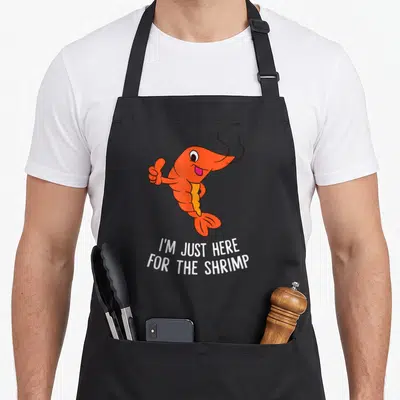 Funny shrimp lover apron - Orange cartoon shrimp thumbs up above white text I'M JUST HERE FOR THE SHRIMP — shown on a black cotton canvas apron against a white background