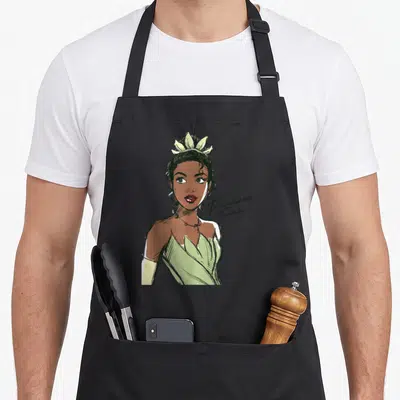 Tiana princess apron - Line art portrait of woman in green dress with Princess Tiana script text on black — shown on a black cotton canvas apron against a white background