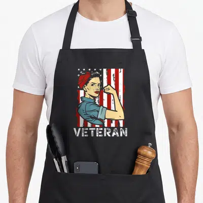 Cute female veteran apron - Rosie the Riveter flexing arm in front of distressed US flag with VETERAN text below — shown on a black cotton canvas apron against a white background
