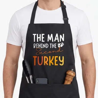 2nd thanksgiving apron - White and orange text reading The Man Behind The Second Turkey with two baby footprints — shown on a black cotton canvas apron against a white background