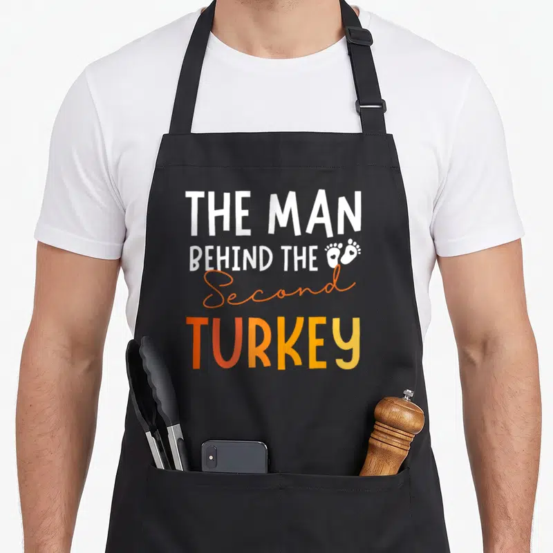 2nd thanksgiving apron - White and orange text reading The Man Behind The Second Turkey with two baby footprints — shown on a black cotton canvas apron against a white background