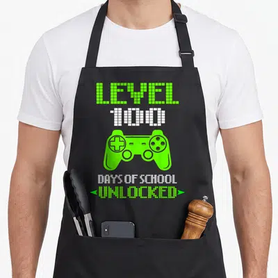 100th video game apron - Pixel art controller with LEVEL 100 DAYS OF SCHOOL UNLOCKED text in lime green and white — shown on a black cotton canvas apron against a white background