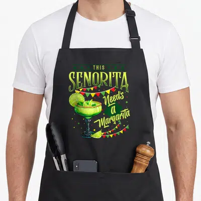 Senorita mayo apron - Cartoon margarita glass with limes and bunting flags under the text This Senorita Needs — shown on a black cotton canvas apron against a white background