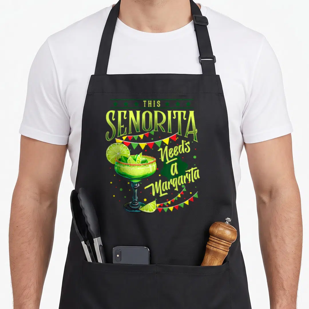 Senorita mayo apron - Cartoon margarita glass with limes and bunting flags under the text This Senorita Needs — shown on a black cotton canvas apron against a white background