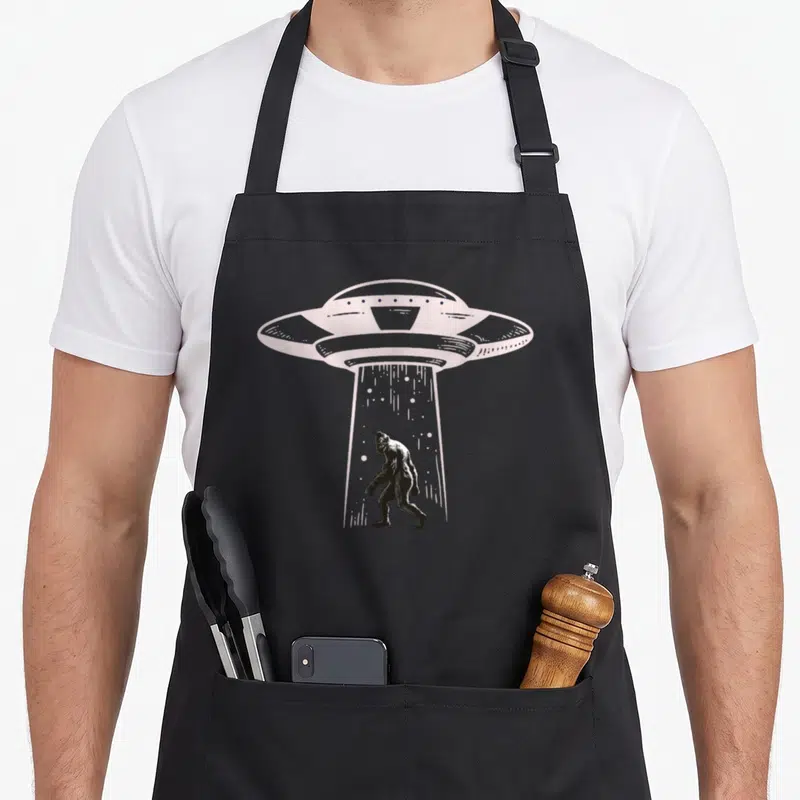 Ufo yeti apron - Line-art UFO emitting beam around walking Bigfoot in grey tones — shown on a black cotton canvas apron against a white background