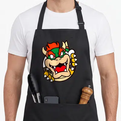 Koopa holiday apron - Cartoon turtle monster head in green yellow red and white with open mouth and claws — shown on a black cotton canvas apron against a white background